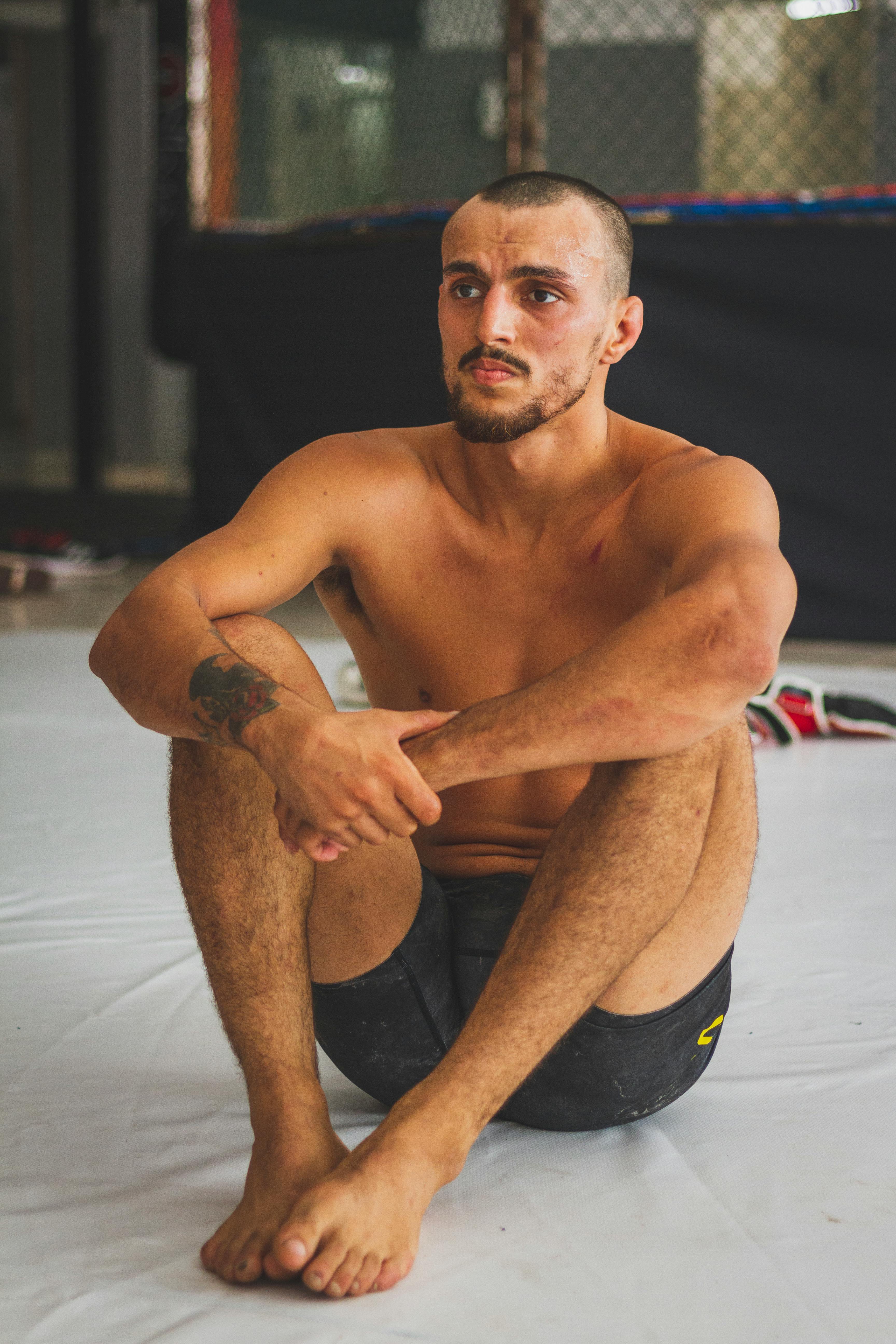 Shirtless MMA fighter sitting on gym floor, reflecting after training session.