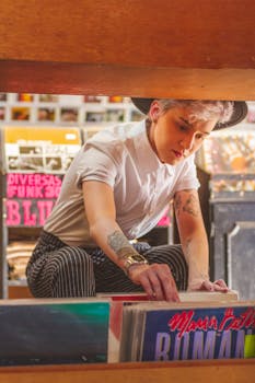 A tattooed adult browsing vinyl records in a music store, wearing a hat and striped pants.