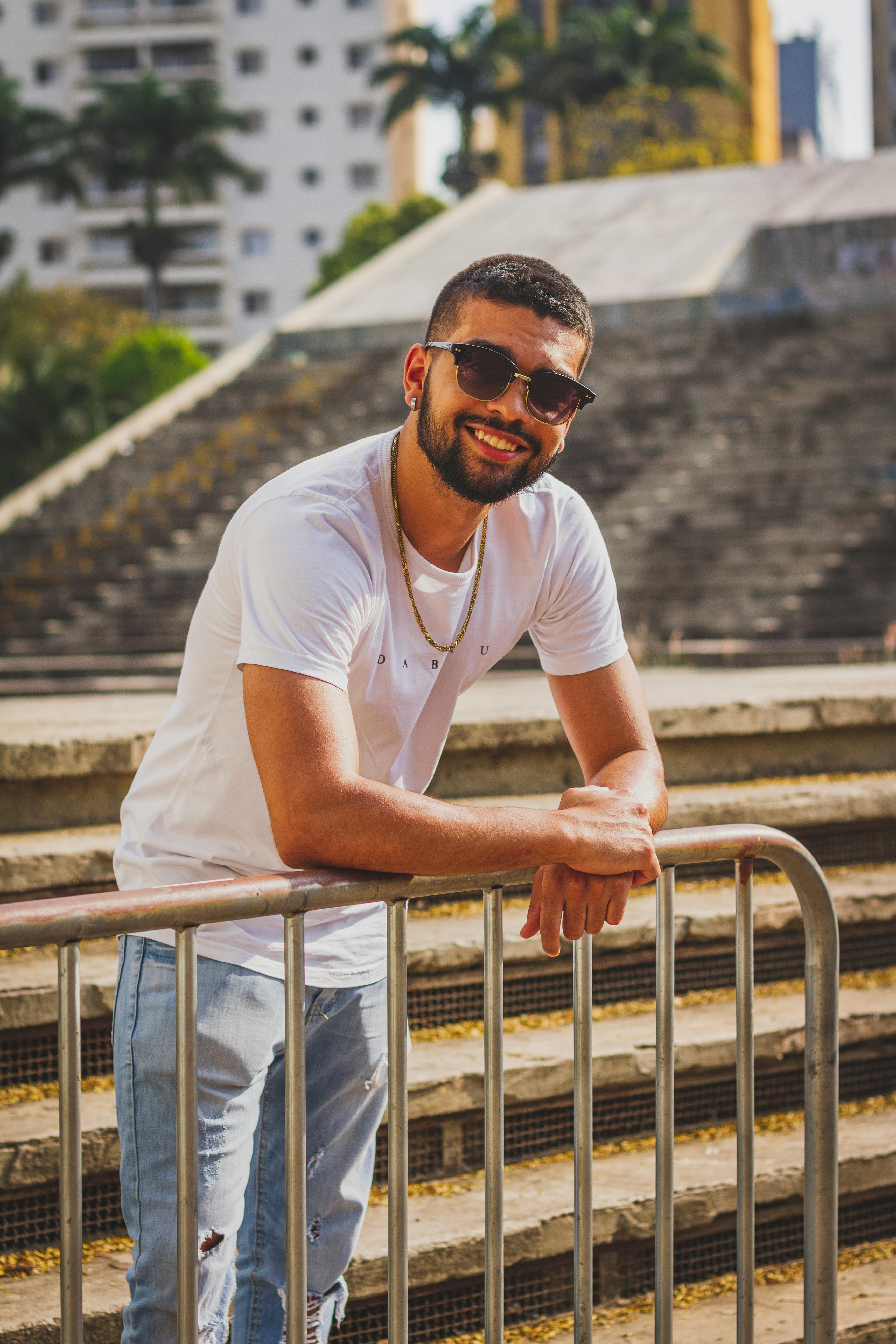 Smiling man in sunglasses and casual attire leaning on railing outdoors.