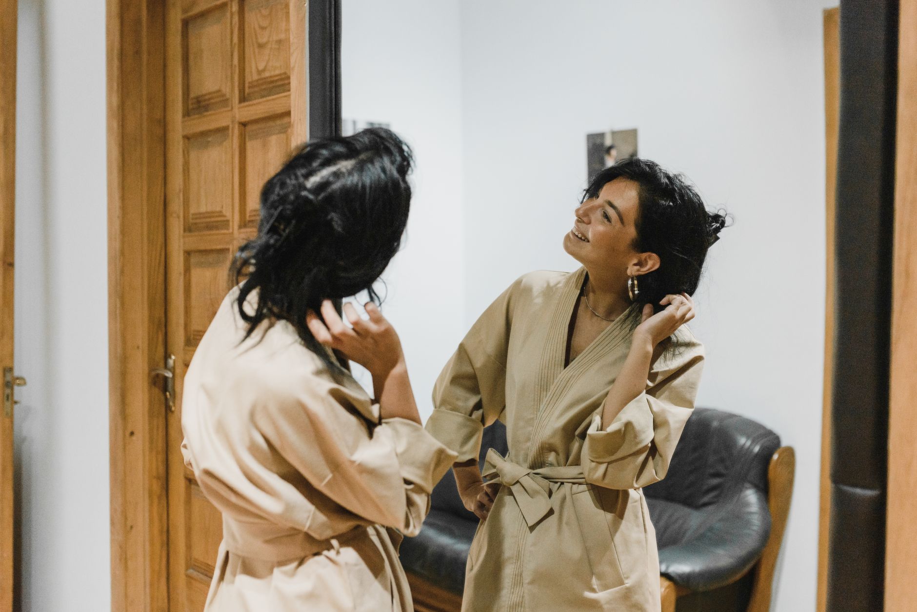A woman in a beige robe smiles into a mirror indoors, creating a serene and intimate moment.