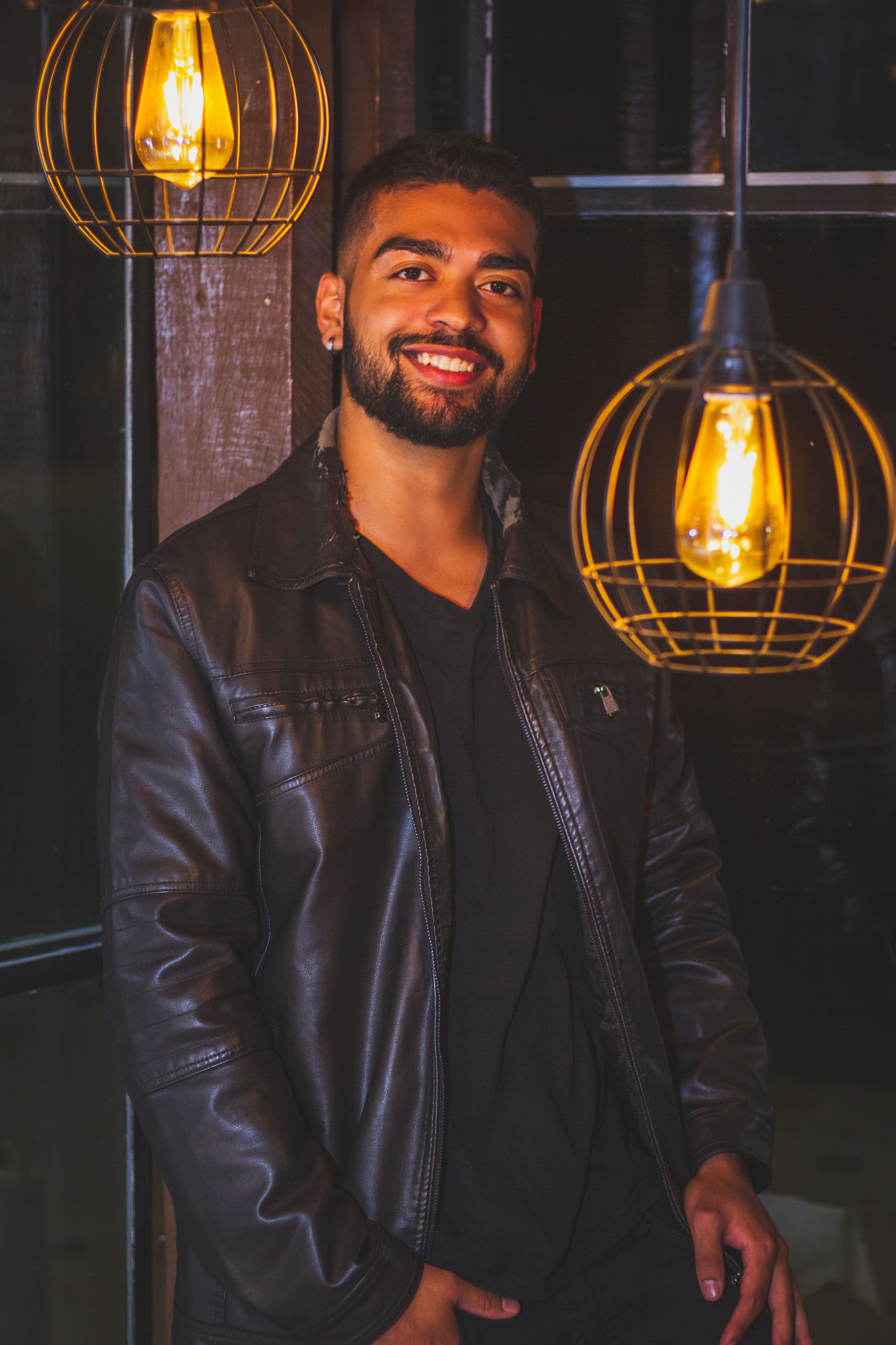 Smiling man in a leather jacket posing indoors with pendant lights.