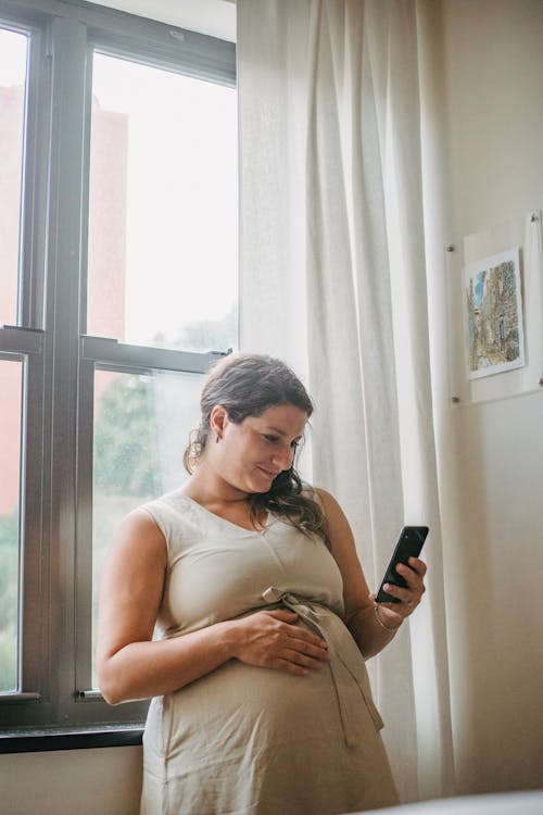 Free Pregnant lady using phone while standing in room Stock Photo