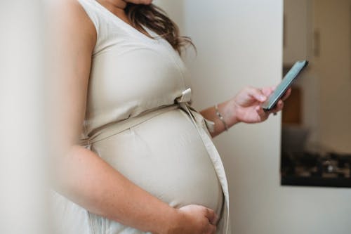 Free Side view crop unrecognizable pregnant woman text messaging on phone while standing in room in daylight Stock Photo