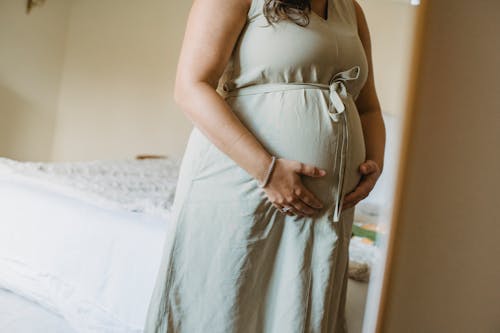 Faceless crop adult pregnant female standing near mirror in room while touching stomach in daytime