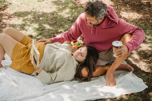 High angle of pregnant woman reading interesting book while spending time with man on picnic on green grass in sunny day