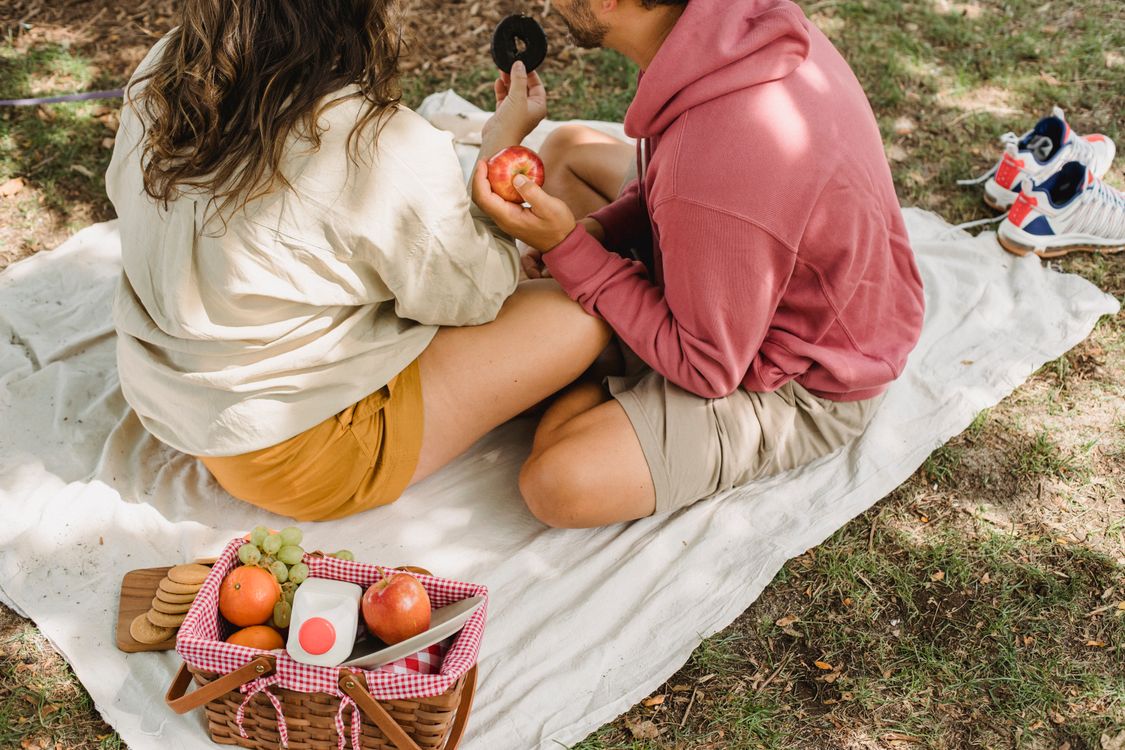 From above of unrecognizable woman and husband with apple sitting on blanket on grass with fruits in wicker basket in countryside