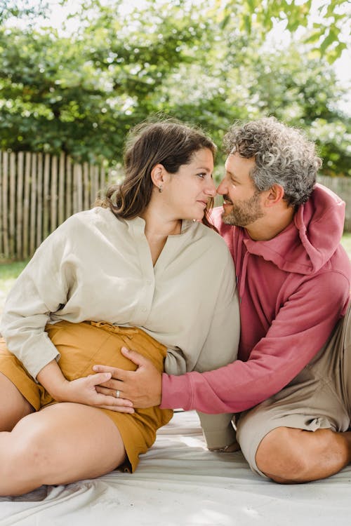 Romantic husband touching belly of pregnant wife while sitting on blanket and looking at each other in countryside during weekend