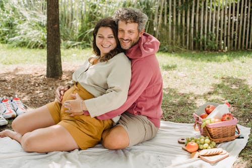 Full body of loving pregnant woman and man embracing while sitting on plaid with food and looking away during romantic moment