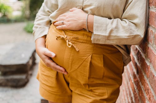 Crop unrecognizable pregnant female in casual outfit standing near brick wall and touching tummy