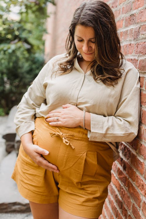 Free Cheerful young pregnant female in casual clothes leaning on shabby brick wall and touching belly Stock Photo