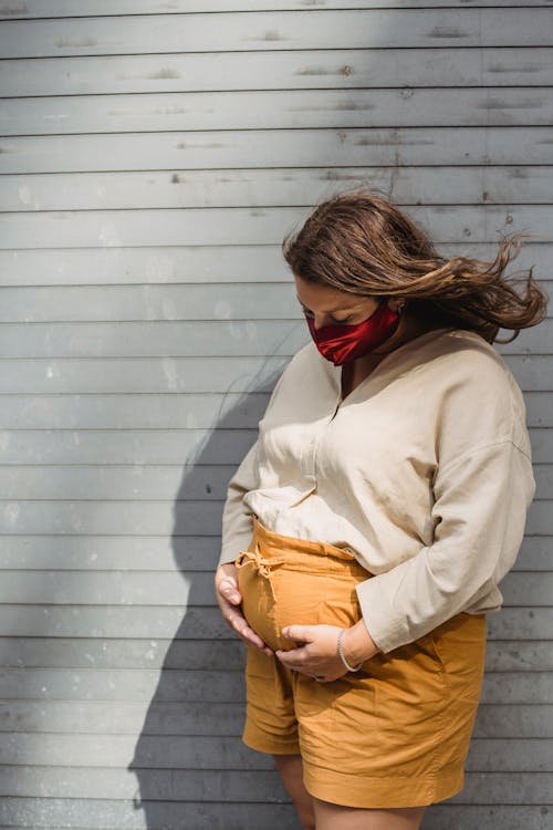 Young pregnant woman in mask and casual outfit touching belly while standing near wall