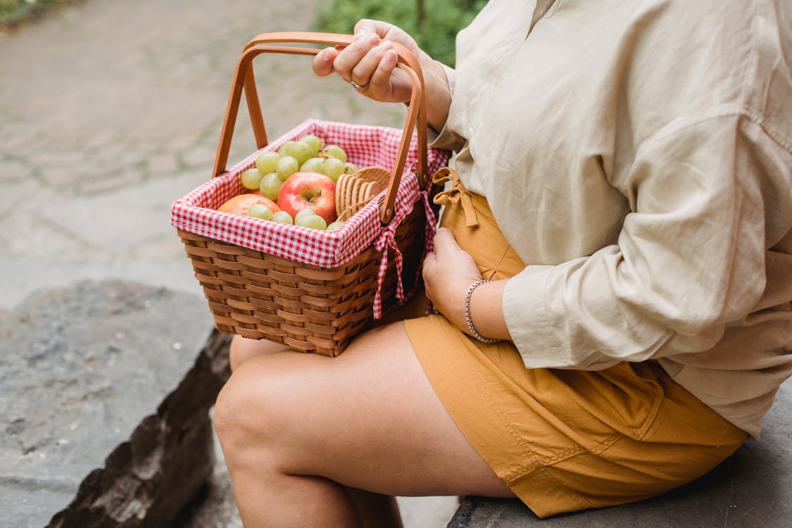 Free Crop anonymous female in expectancy wearing casual clothes with basket full of ripe fruits and biscuits prepared for picnic Stock Photo