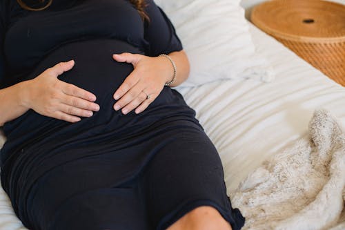 From above of crop anonymous expectant female in dress stroking tummy on cozy bed at home