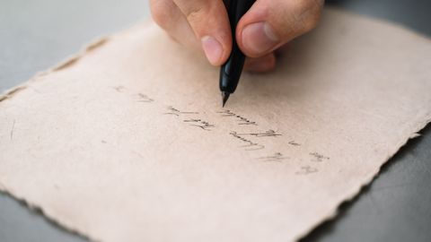 A close-up view of a person's hand writing with a black pen on textured paper.