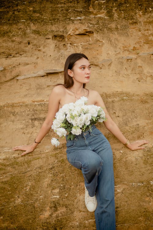 Free Calm young female with bare skin covering breast with bouquet of roses while leaning on rough formation Stock Photo