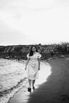 Black and white of trendy barefoot ethnic female adolescent strolling on sandy ocean beach under cloudy sky while looking down