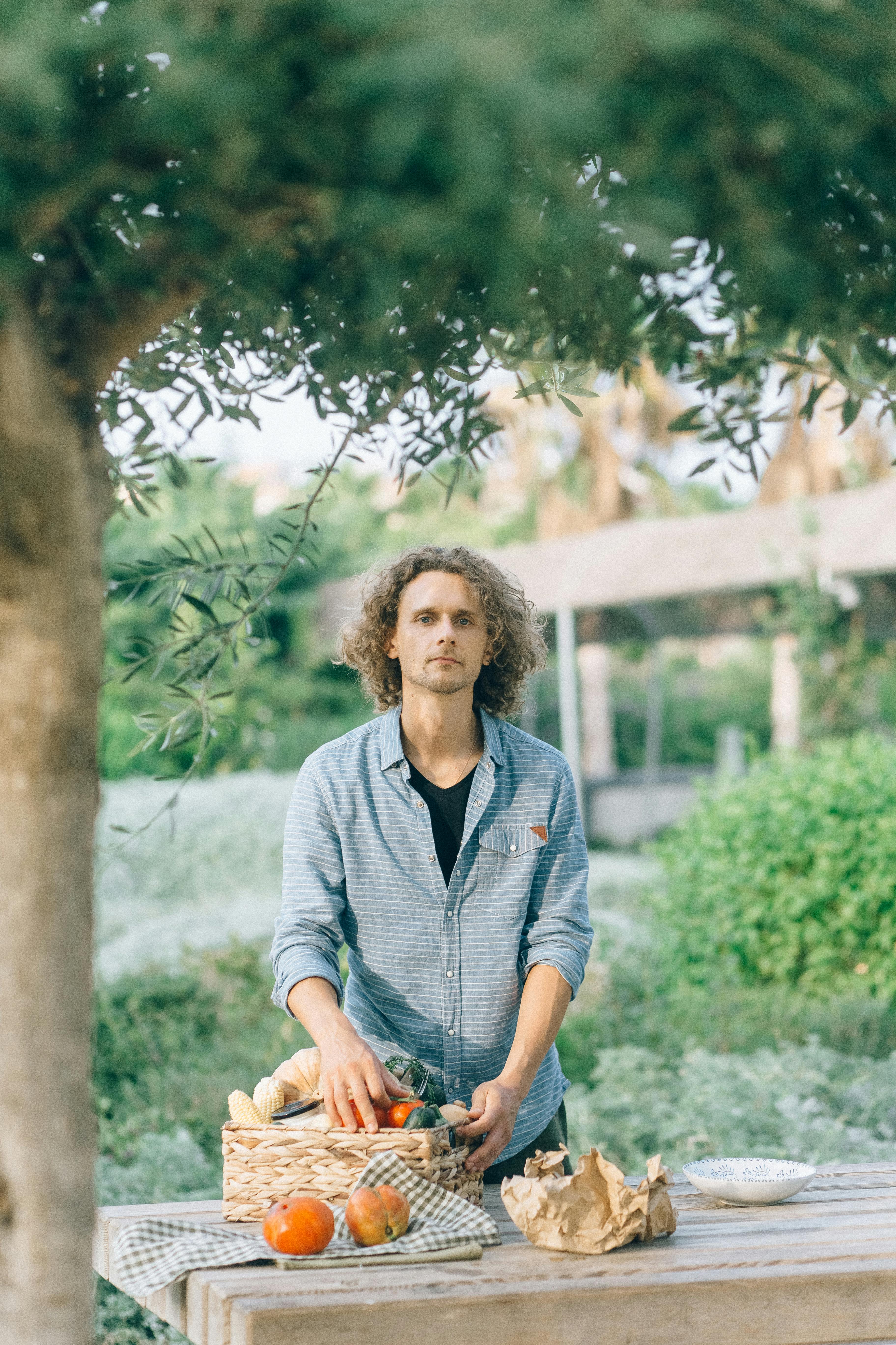 A man outdoors with a woven basket of fresh vegetables, standing at a table under an olive tree.
