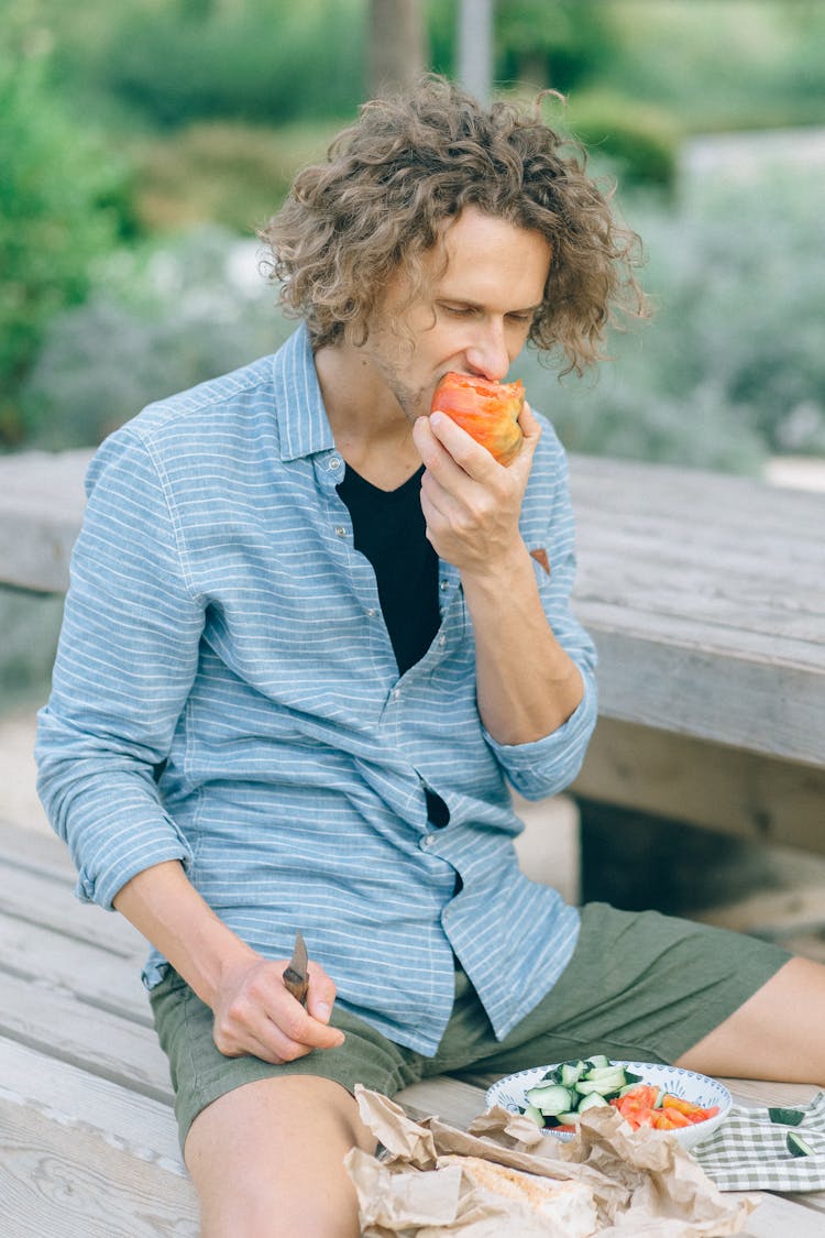 Man Sitting On Wooden Bench Eating A Tomato