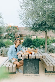 A man sitting at a wooden picnic table outdoors surrounded by greenery.