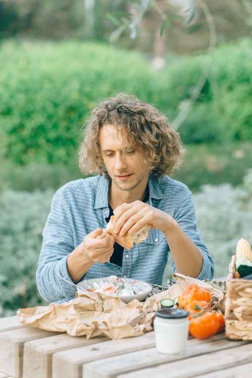 Free Man with curly hair eating a healthy meal outdoors, seated at a rustic picnic table. Stock Photo
