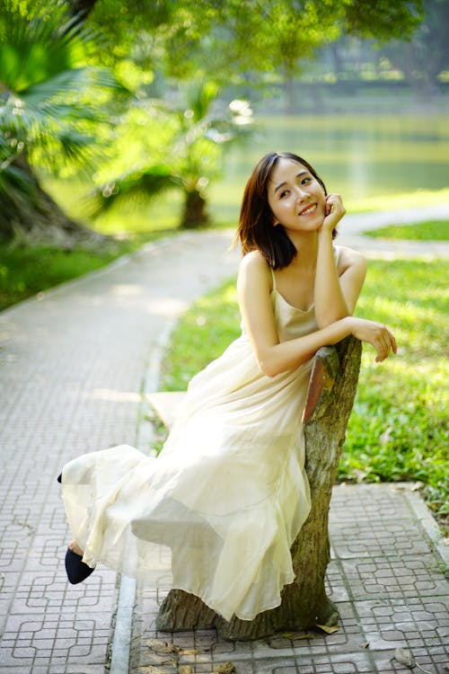 Full body of cheerful Asian female in white dress looking away while sitting on bench in park on summer day
