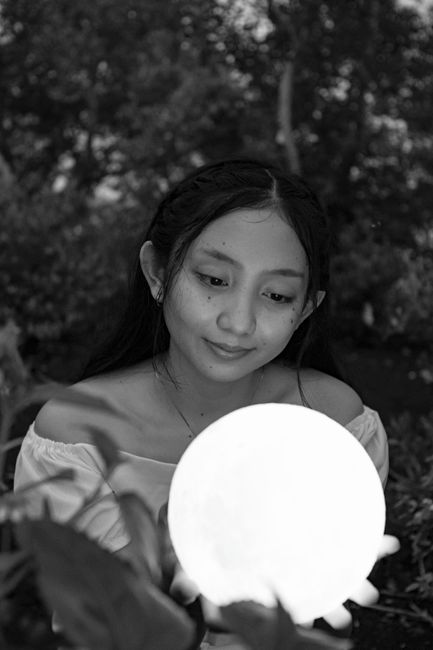 Black and white of glad contemplative ethnic female adolescent with glowing glass ball looking down in garden