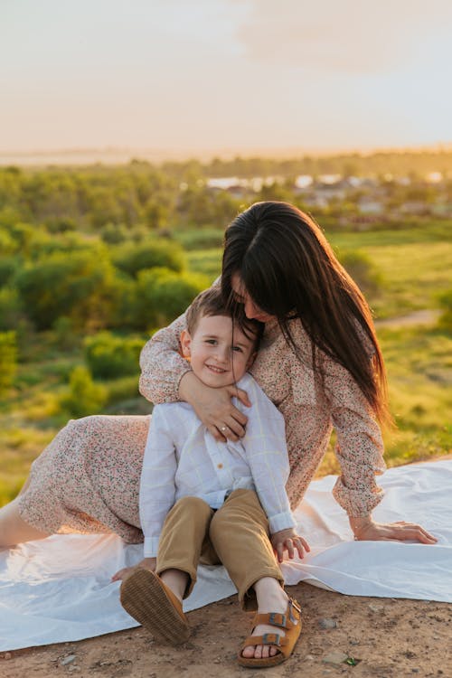 Cheerful mother with long hair hugging preschooler son while sitting on blanket on sandy ground on edge of hill with green trees on background