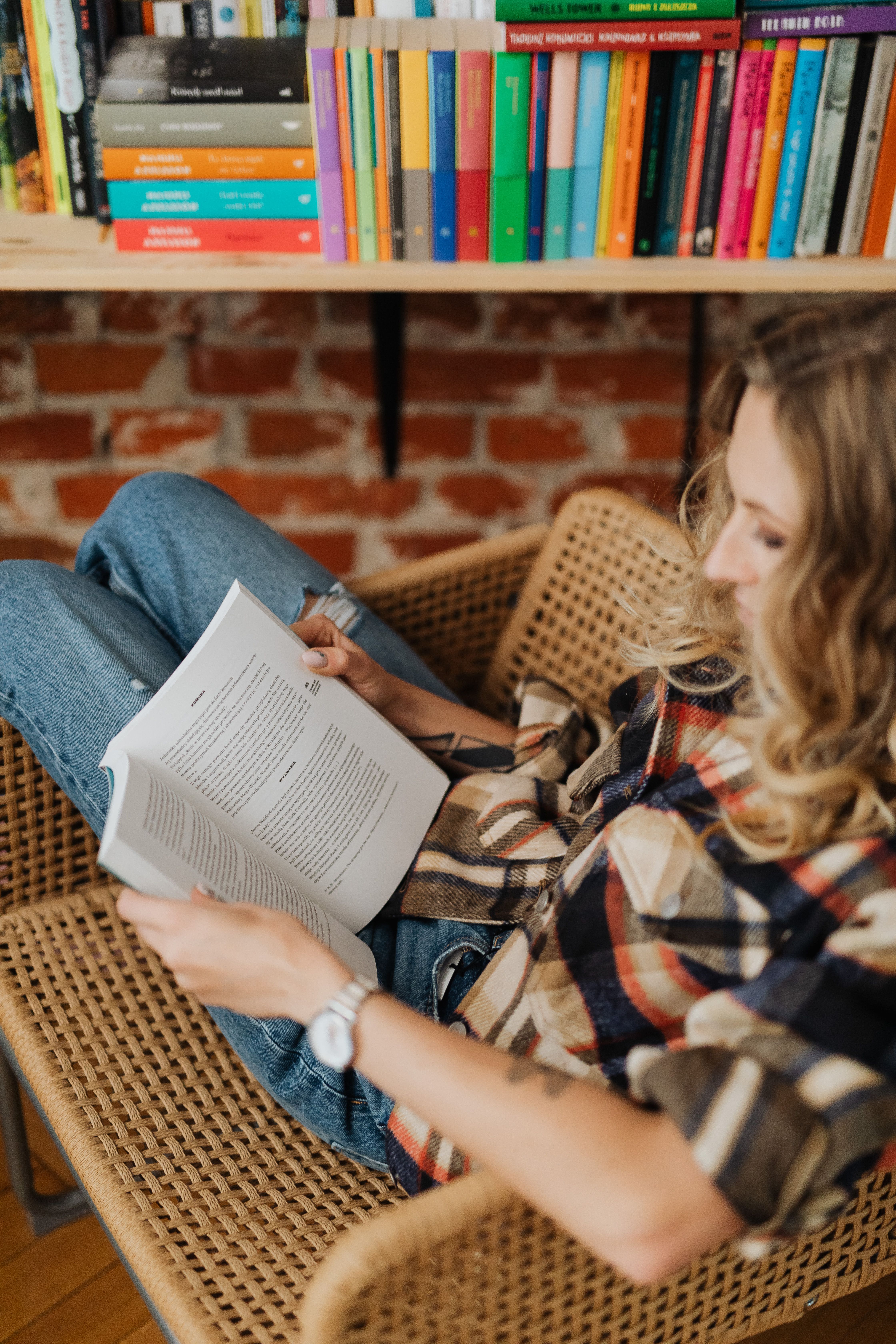 A woman reading in a wicker chair beside a colourful bookshelf