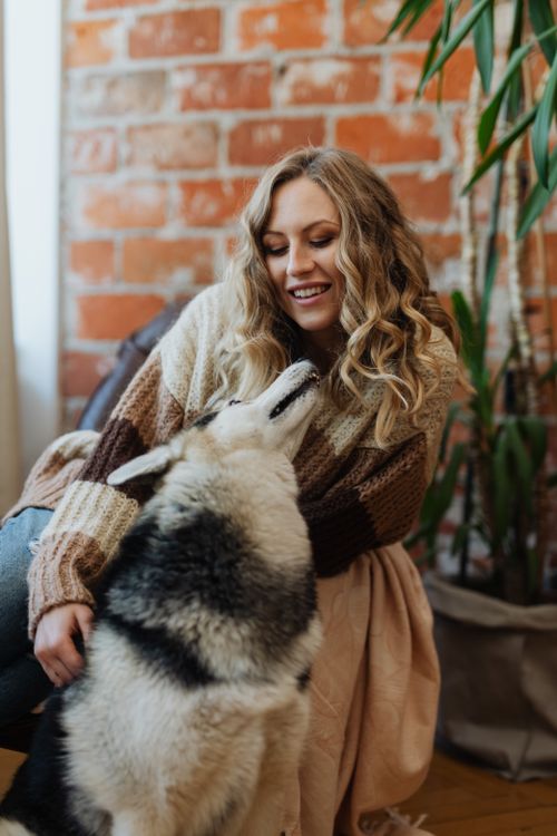 Free A woman shares a joyful moment indoors with her affectionate pet dog, posing against a brick wall. Stock Photo