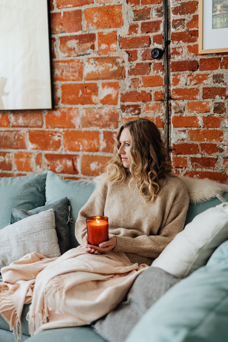 Woman Sitting On A Sofa Wrapped In A Blanket And Holding A Burning Candle 