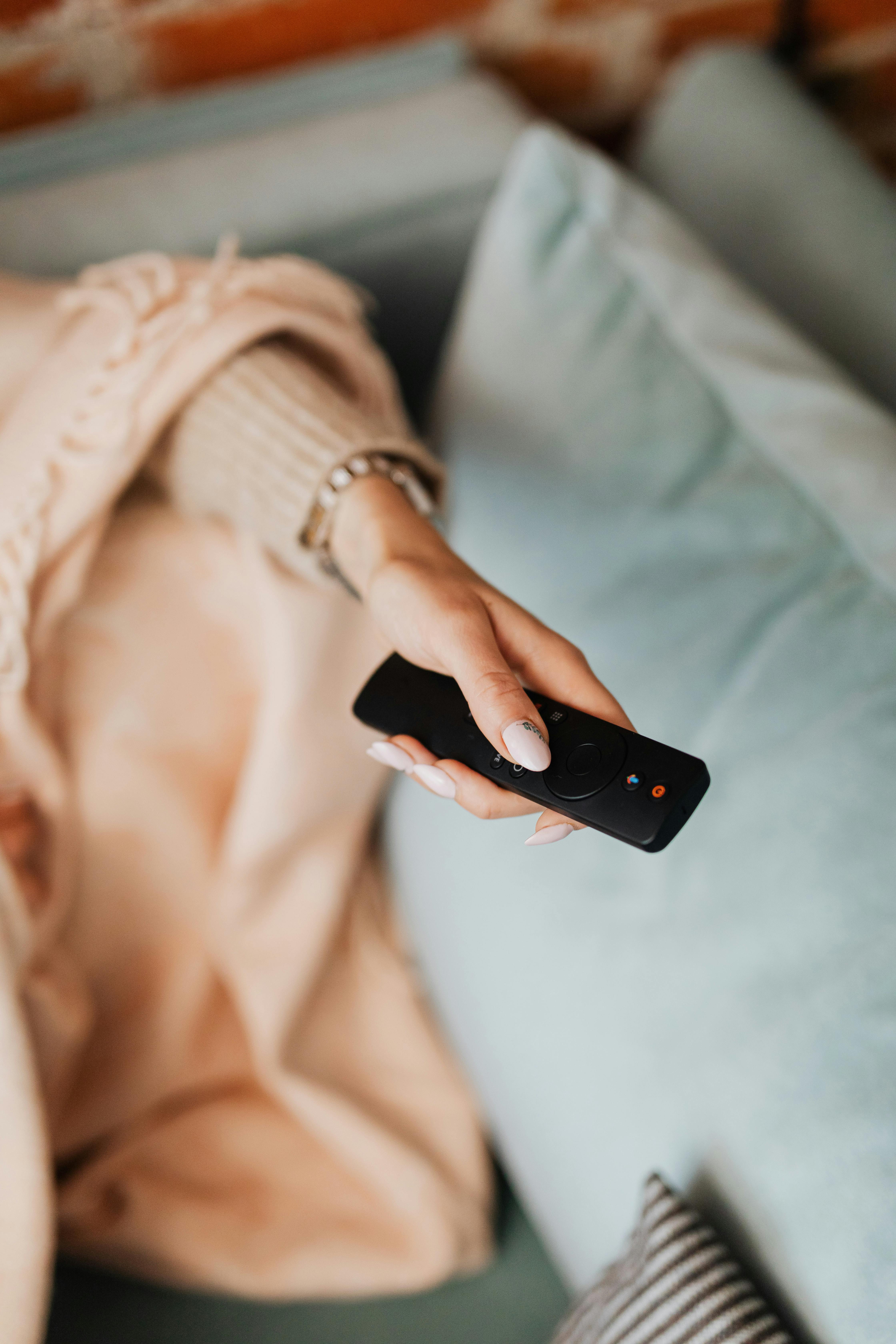 Woman's hand holding a TV remote over a cozy blanket on a couch.