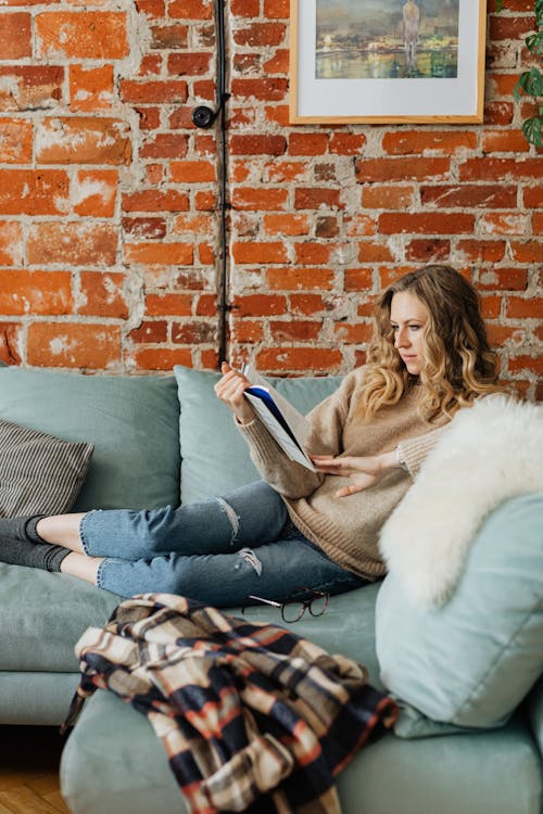 Free Woman in Sweater Sitting on Sofa and Reading a Book Stock Photo
