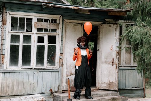 Free A person dressed as a creepy clown stands outside an abandoned house holding a red balloon. Stock Photo