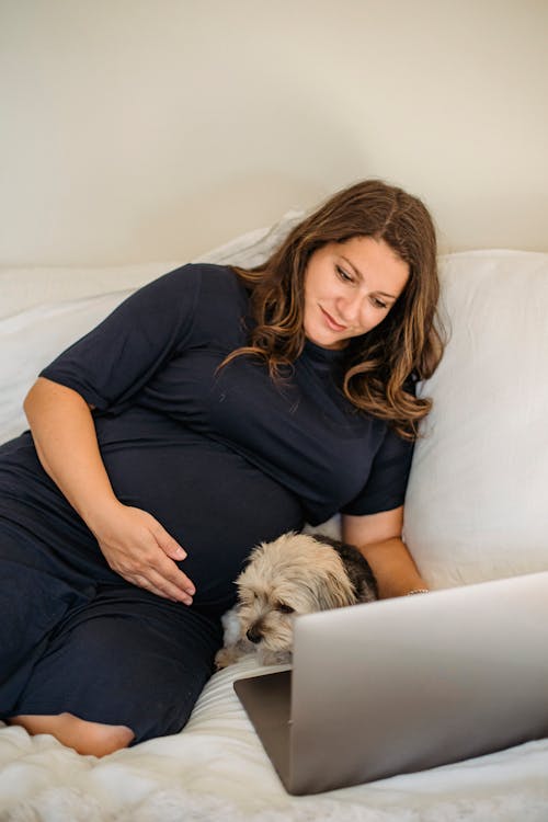 Smiling pregnant woman with dog watching laptop on cozy bed