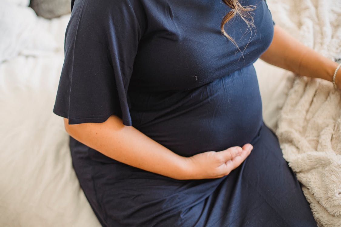 From above of crop anonymous expectant female caressing tummy on cozy bed at home in daylight