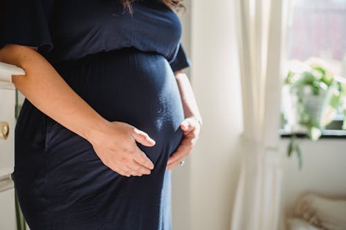 Crop anonymous expectant female in dress stroking tummy in room illuminated by sunlight at home