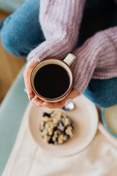 A close-up of hands holding a cup of black coffee, set against a breakfast bowl.