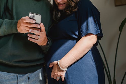 Crop anonymous man showing cellphone to smiling expectant wife while spending time together in house in sunlight