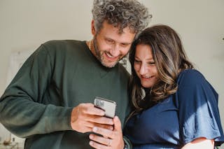 Crop glad unshaven man in casual apparel showing mobile phone to feminine female beloved at home