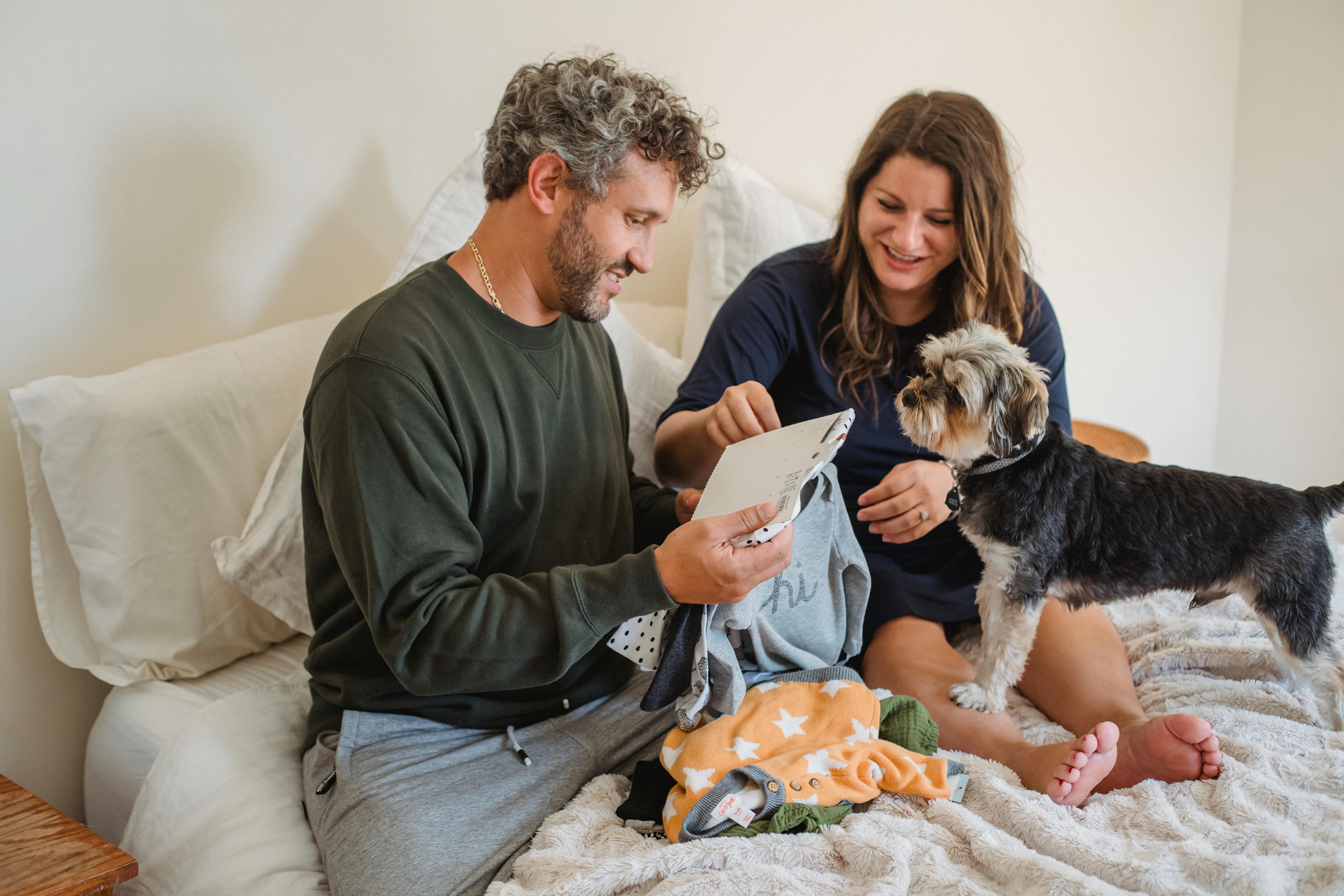 smiling couple with dog and baby clothes resting on bed