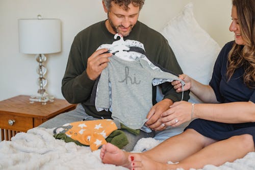 Crop pregnant woman with husband watching baby wear on bed