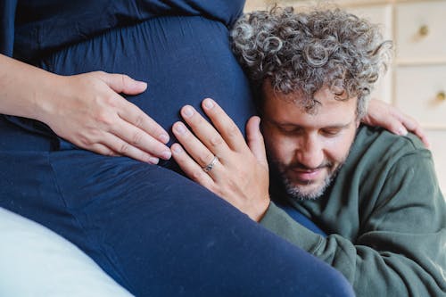 Free Smiling bearded husband with curly gray hair and closed eyes embracing gently tummy of crop unrecognizable pregnant wife at home Stock Photo