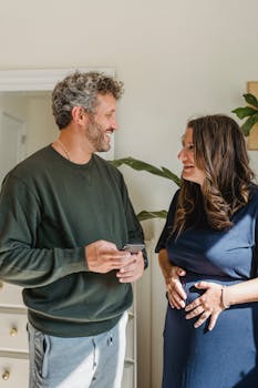 Adult smiling expectant female speaking with bearded male partner with cellphone while looking at each other in house