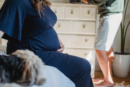 Crop unrecognizable wife touching tummy while sitting on bed against busy husband in apartment