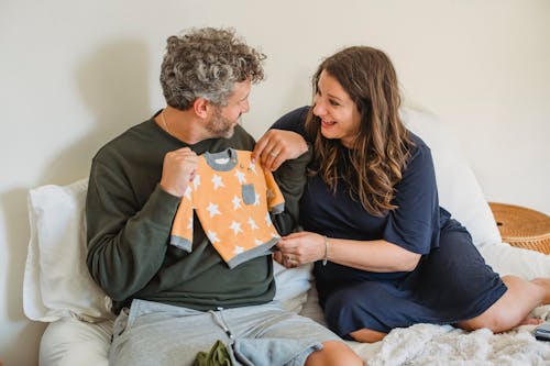 Cheerful pregnant couple resting on bed while preparing clothes for infant and looking at each other