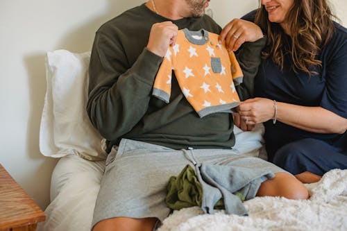 Crop faceless couple considering new apparel for newborn baby while resting on soft bed