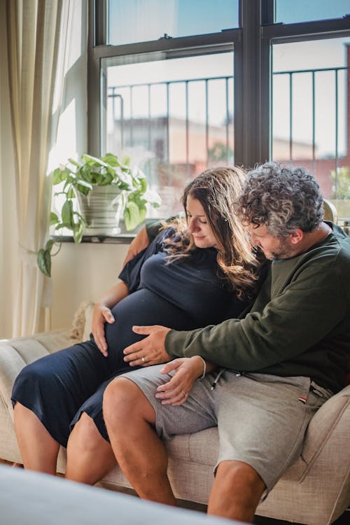 Cheerful adult man wearing shorts resting with pregnant wife on couch near window in light apartment while gently touching belly