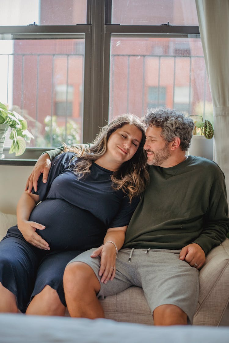 Positive Husband Hugging Pregnant Wife While Sitting On Sofa