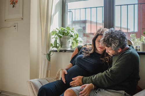 Calm adult woman looking at husband while cuddling gently and feeling baby in pregnant belly while chilling on cozy sofa in light room