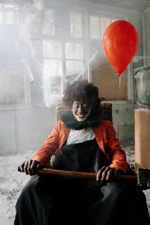 Free A creepy clown sits with a hammer in an eerie abandoned building, holding a red balloon. Stock Photo